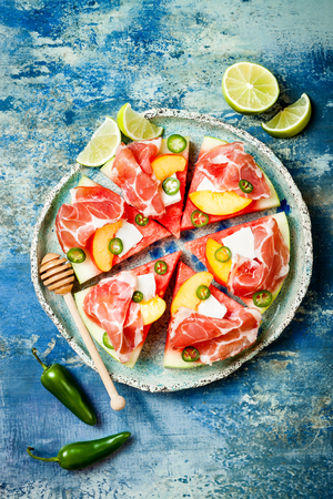 Fresh summer watermelon pizza with feta cheese, peach, prosciutto, jalapeno and honey drizzle on blue backgroundの写真素材