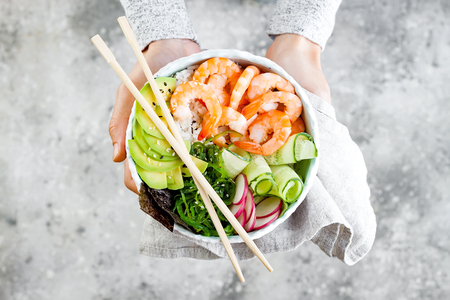 Girl holding shrimp poke bowl with seaweed, avocado, cucumber, radish, sesame seeds.の写真素材
