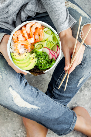 Girl in jeans holding shrimp poke bowl with seaweed, avocado, cucumber, radish, sesame seeds.の写真素材