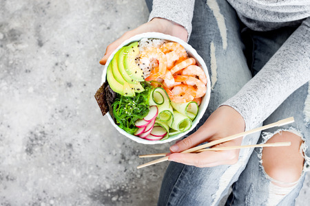 Girl in jeans holding shrimp poke bowl with seaweed, avocado, cucumber, radish, sesame seeds.の写真素材