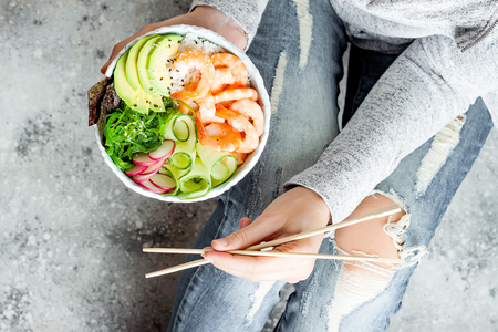 Girl in jeans holding shrimp poke bowl with seaweed, avocado, cucumber, radish, sesame seeds.の写真素材