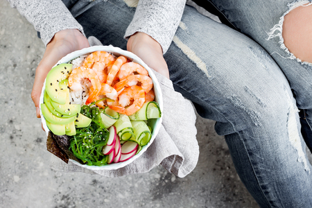 Girl in jeans holding shrimp poke bowl with seaweed, avocado, cucumber, radish, sesame seeds.の写真素材