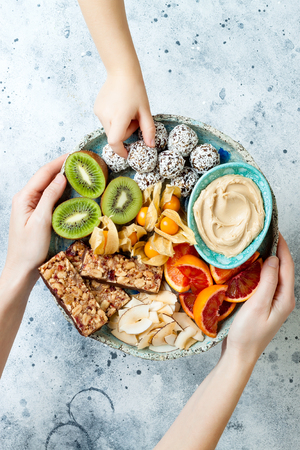 Mother giving healthy vegan dessert snacks to toddler child. Concept of healthy sweets for children. Protein granola bars, homemade raw energy balls, cashew butter, toasted coconut chips, fruits platterの写真素材
