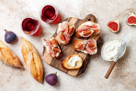 Crostini with prosciutto, cream cheese and figs on wooden board. Appetizers, antipasti snacks and red wine in glasses. Authentic traditional spanish tapas set. Light beige background. Top viewの写真素材