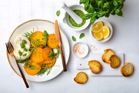 Persimmon caprese salad with burrata cheese, arugula, pesto and toasted bread. Winter salad recipe. Top view, overhead, flat layの写真素材