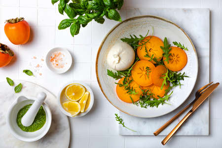 Persimmon caprese salad with burrata cheese, arugula, pesto and toasted bread. Winter salad recipe. Top view, overhead, flat layの写真素材