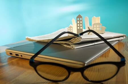 Magnifying glass in front of an open newspaper with paper houses. Concept of rent, search, purchase...