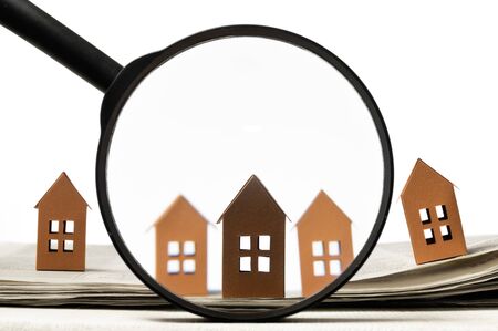Magnifier in front of an open newspaper with paper houses on white background. Concept of rent,...