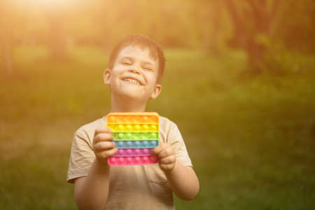 Gigglet Boy Has Fun With His Colorful and Sunbathes in the Bright Summer Sun. Young One Smiling for a Sun in the Summer Square. Trees and Grass Background. High quality photoの写真素材