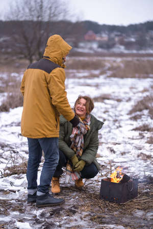 Mature couple at camp fire, happy family on winter vacation. away in wild. Smiling middle aged man and woman in snowy nature or park together. Family seasonal vacation concept.の写真素材