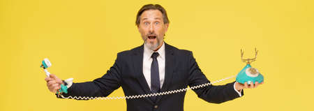 Amazed, shocked middle aged businessman in black suit with old fashion telephone in hands stretched...