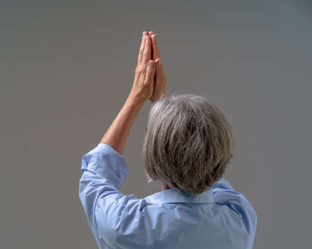 Senior woman with grey hair praying staying back to the camera isolated on greying background. Begging god for help woman put hands together lifted high wearing blue shirt. Online with God.の写真素材