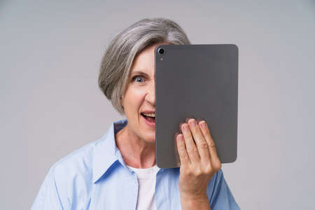 Angry mature grey haired woman hide behind digital tablet posting bad comments or trolling other users online in social media. Senior woman in blue shirt isolated on white background.の写真素材