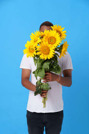 Hide behind bouquet of sunflowers young African American man wearing white t-shirt and isolated on blue background. Romantic young man with flowers. Man with summer flowers. No face visible.の写真素材