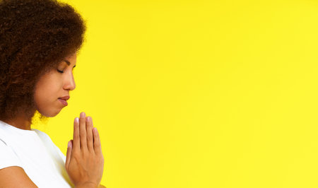 Young african american woman in prayer position wearing white t-shirt isolated on yellow background. African girl praying God studio shot. Spiritual woman put hands in prayer.の写真素材