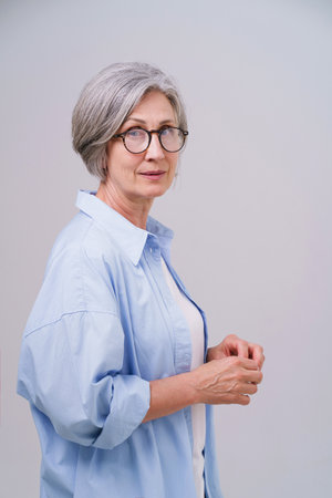 Mature gray haired beautiful businesswoman wearing long blue shirt stands sideways with hands together looking straight in camera isolated on white background. Mature people beauty concept.の写真素材