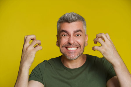 Middle aged caucasian gray haired man showing anger facial expression with hands up wearing khaki t-shirt isolated on yellow background. Handsome man showing his grin with perfect teeth.の写真素材