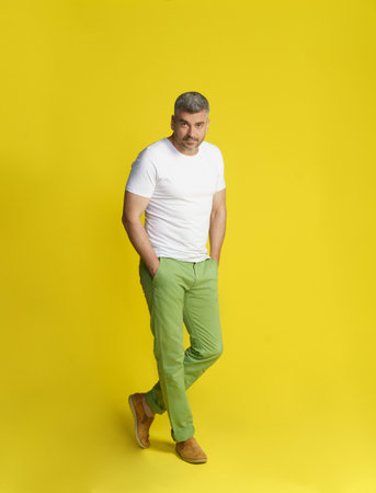 Middle aged handsome gray hair man posing with hands in pockets and crossed legs wearing white t-shirt and green jeans isolated on yellow background. Fit, muscular middle aged man.の写真素材