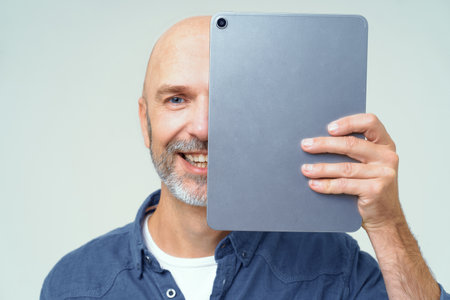 Handsome bold man cover half of his face with digital tablet in hand smiling looking at camera isolated on white background. Happy freelancer man with tablet. Using modern technologies bald man.の写真素材