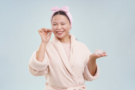 Asian woman wearing a bathrobe, smiling while smelling cosmetic cream on toned background. Sense of joy, happiness, and self-care, highlighting the importance of taking care of oneself and feeling good about it. High quality photoの写真素材