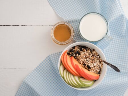 Healthy and tasty breakfast - muesli, slices of persimmon and apple, a glass of fresh milk and maple syrup in a cup on a white table topの写真素材