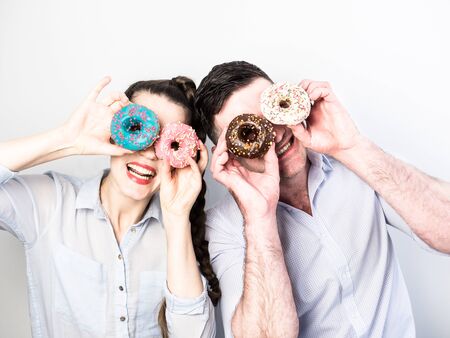 Funny and in love couple with colorful donuts on a background a white wall. Pink, blue, white and brown colors.の写真素材