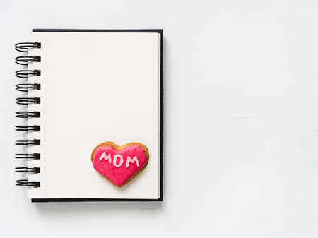 Fresh delicious cookies in the shape of a heart with the word MOM, covered with glaze, an album with a blank page for Your inscription.White isolated background.Top view.Concept of a happy familyの写真素材