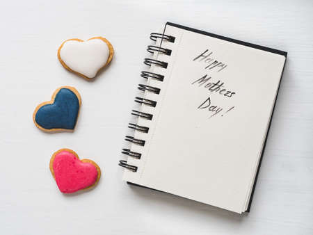 Fresh, fragrant cookies in the shape of a heart, covered with multicolored glaze, sketchbook with a beautiful, caligraphic inscription about congratulation on Mother's Day.Top view,white backgroundの写真素材