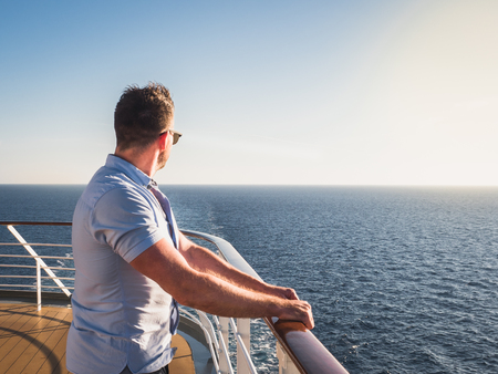 Attractive man in sunglasses on the top deck of a cruise ship looking out into the distance against the background of a sunset. Concept of sea travel and recreationの写真素材