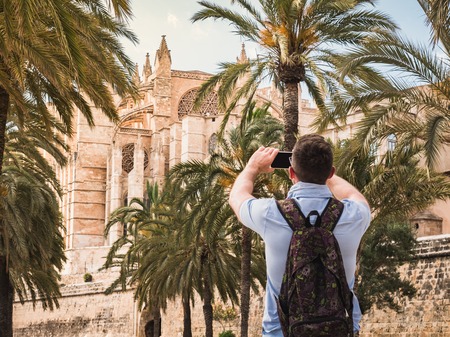 Stylish, happy man with a smartphone on the background of a river, palm trees and historic, famous building in a summer, sunny dayの写真素材