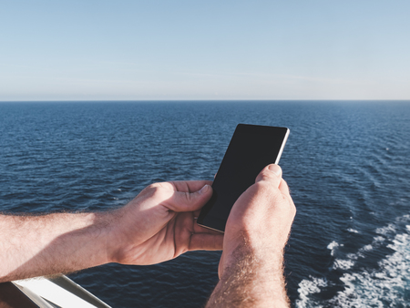 Men's hands holding a mobile phone on the deck of a cruise ship against the background of sea wavesの写真素材