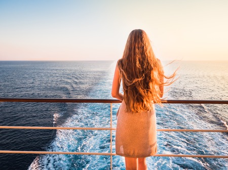 Stylish, young woman on an empty deck of a cruise ship against a background of sea waves, blue sky and sunsetの写真素材
