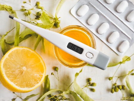Electric thermometer, pills, fresh yellow lemon, jar of honey, linden leaves and flowers on a white,...