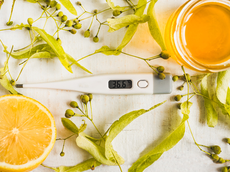 Electric thermometer, pills, fresh yellow lemon, jar of honey, linden leaves and flowers on a white,...