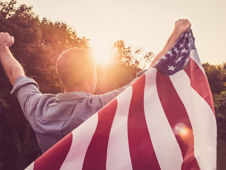 Attractive man holding a US flag against the background of the rays of the setting sun. View from the back. Preparing for the holidaysの写真素材
