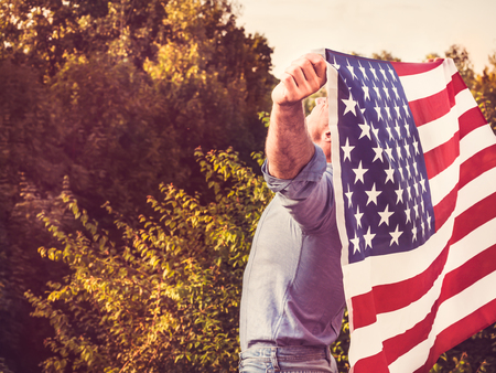 Attractive man holding waving in the wind US flag against the backdrop of the rays of the setting...