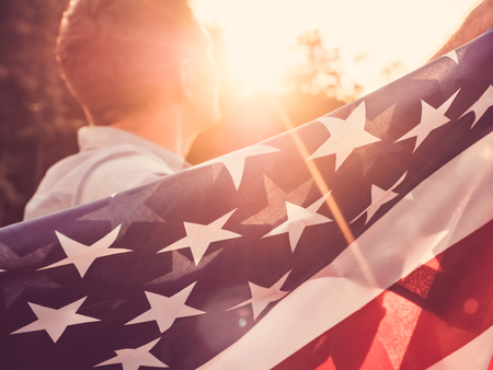 Attractive man holding waving in the wind US flag against the backdrop of the rays of the setting sun. View from the back, closeup. Preparing for the holidaysの写真素材