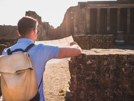 Handsome man in sunglasses looking at a map with landmarks against the backdrop of historic ruins, blue sky and the rays of the setting sun. Italy, Pompeii. Travel and vacation conceptの写真素材