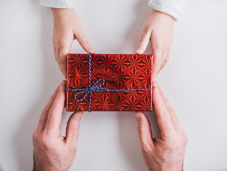 Bright box with a gift, tied with a ribbon, hands of a loving father and hands of a child on a white, isolated background. Top view, close-up. Preparing for the holidays. Happy family conceptの写真素材
