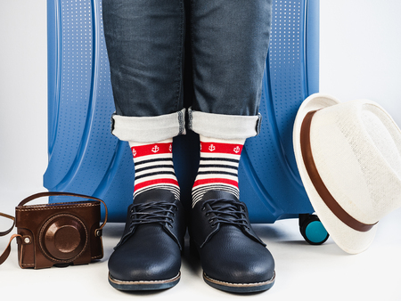 Stylish suitcase, men's legs, vintage camera, sunhat, multicolored socks with a nautical theme and shoes on a white, isolated background. Close-up. Concept of style, fashion, beauty and travelの写真素材