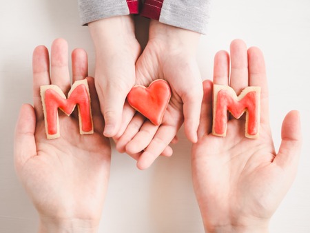 Hands of a younger daughter and her mother's, beautiful glazed cookies and word MOM on a white, isolated background. Top view, close-up. Preparing for the holidaysの写真素材