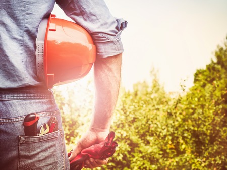 Handsome man in work clothes with tools, standing on a background of trees, blue sky and and rays of...