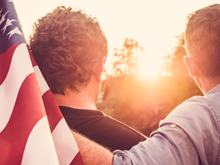 Two friends holding an American Flag against a background of trees and blue sky. View from the back, close-up. National holiday conceptの写真素材