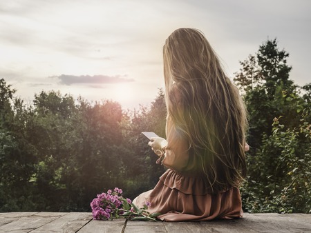 Stylish woman with a mobile phone in a beautiful dress sitting on a wooden terrace against the sky, the setting sun and trees. Concept of rest and beautyの写真素材