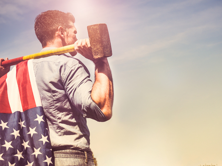 Attractive man holding a sledgehammer and a US Flag in his hands and looking into the distance against a background of trees, blue sky and the rays of the setting sun. National holiday conceptの写真素材