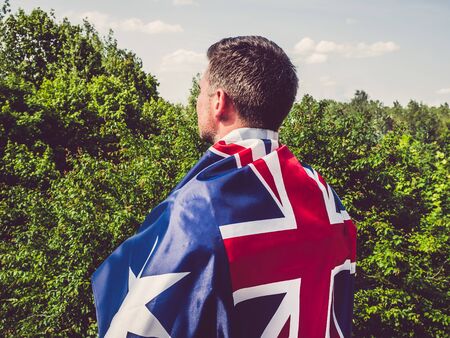 Handsome man waving the Flag of Australia against the backdrop of trees and blue sky. View from the back, close-up. National holiday conceptの写真素材