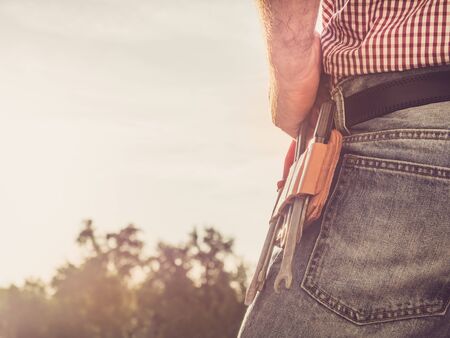 Male hand, blue jeans and vintage belt with tools in the park against the backdrop of green trees and of the rays the setting sun. Close-up. Concept of labor and employmentの写真素材