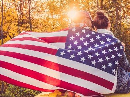 Happy married couple holding the US flag against the background of yellow trees and the setting sun. Happy relationship conceptの写真素材