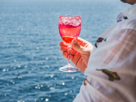 Fashionable man holding a beautiful glass of pink wine on the open deck of a cruise liner against the backdrop of blue sea waves. Side view, close-up. Concept of leisure and travelの写真素材