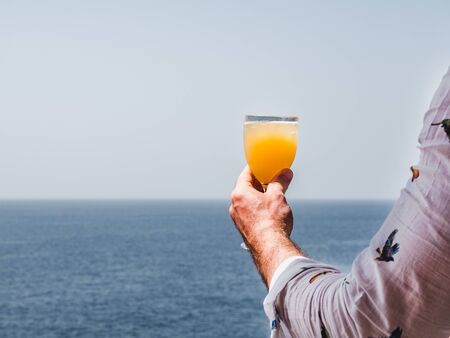 Fashionable man holding a beautiful glass of wine on the open deck of a cruise liner against the backdrop of blue sea waves. Side view, close-up. Concept of leisure and travelの写真素材